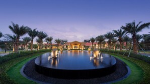 Un hotel con una fuente central, palmeras y un césped bien cuidado.