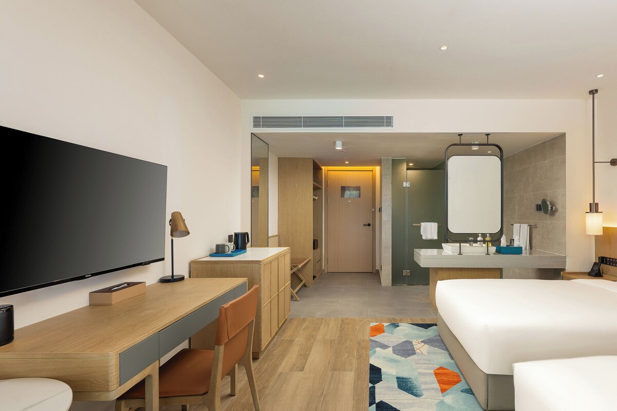 Een hotelkamer met een bed, een bureau met stoel, een televisie, een badkamer met spiegel en een douche.