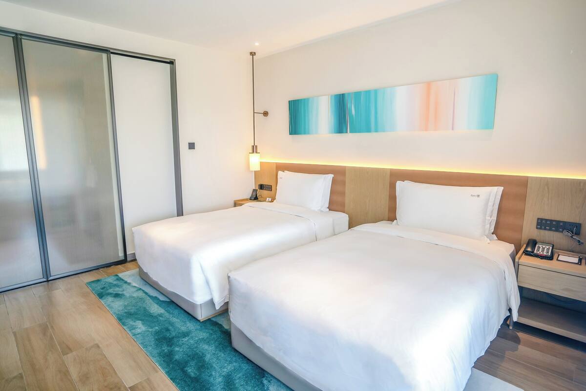 Habitación de hotel con dos camas, cabecero de madera, mesita de noche con lámpara, teléfono y un cuadro en la pared.