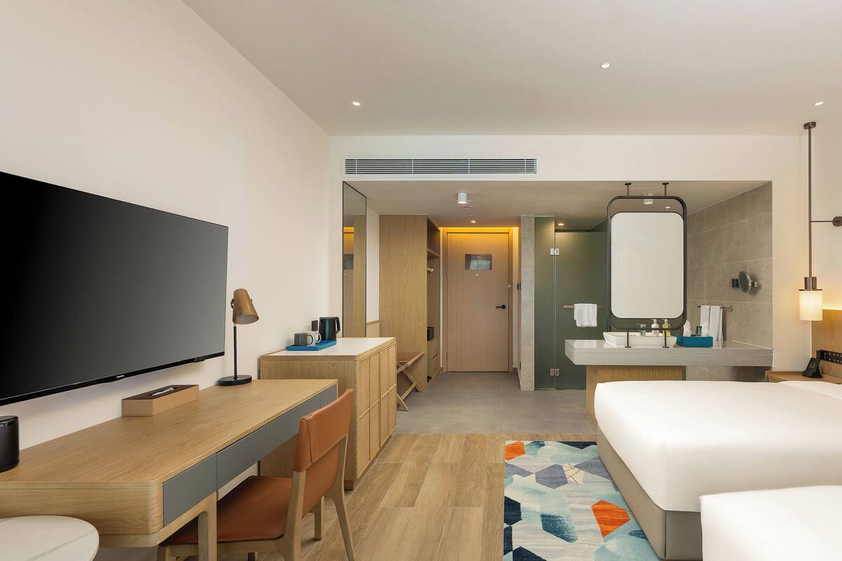 Una habitación de hotel con cama, escritorio con silla, televisor, baño con espejo y ducha.