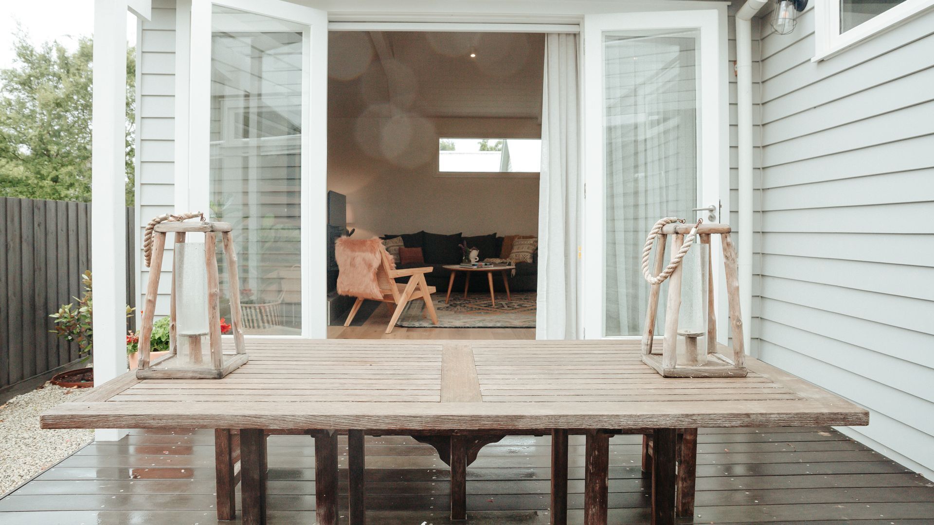 Une terrasse en bois avec une table et deux lanternes, qui mène à une porte ouverte donnant sur un salon équipé d’un canapé et d’une table basse.