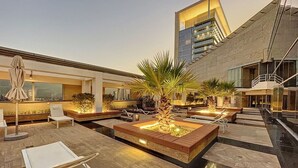 A rooftop terrace with outdoor seating, palm trees, and a modern building in the background.