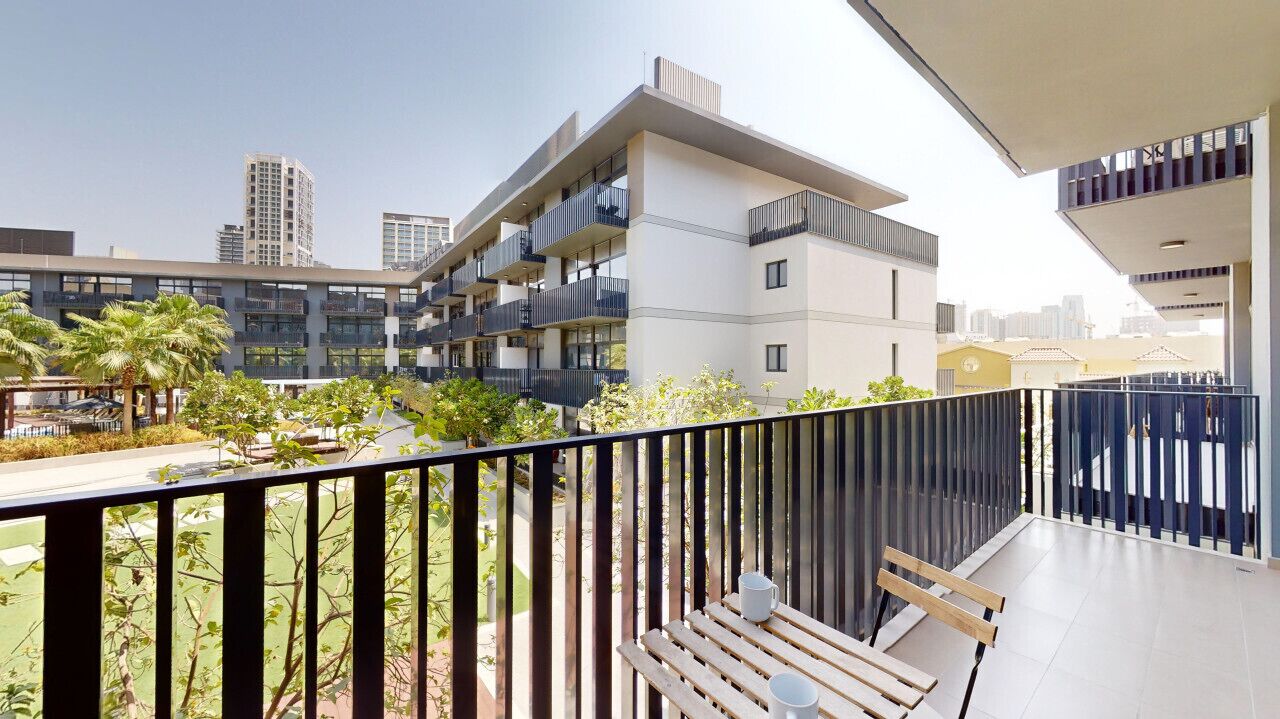 A modern apartment building with balconies, a wooden bench, and a view of palm trees and other greenery.
