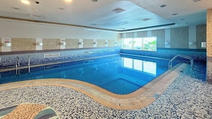 Uma grande piscina interior com piso e paredes revestidas, rodeada por uma área de lazer com assentos e corrimão.