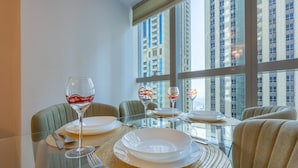 Mesa de jantar posta para quatro pessoas, com copos e pratos transparentes, localizada em um ambiente com grandes janelas com vista para um horizonte urbano.
