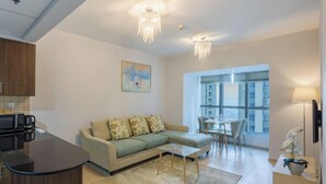 Uma sala de estar moderna com sofá, mesa de centro e área de jantar. Há uma cozinha com micro-ondas e uma mesa de jantar com cadeiras. O quarto possui grandes janelas e um candelabro.