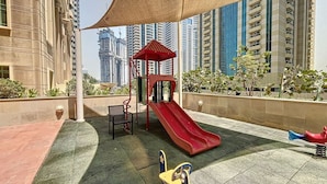 Um parquinho no terraço com um escorrega vermelho, um abrigo vermelho pequeno e um conjunto de balanço amarelo.