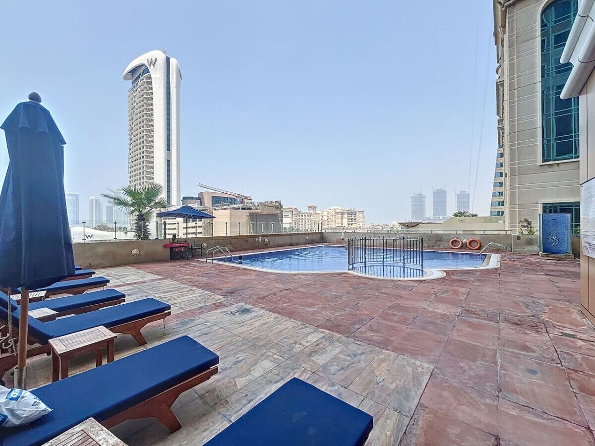 Zona de piscina en la azotea con sillas de descanso y sombrillas, rodeada de edificios de la ciudad.