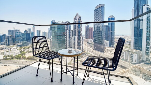 A balcony with two chairs and a small table, overlooking a cityscape with modern skyscrapers. - 2bedroom Apt in Burj Crown (Dubai)