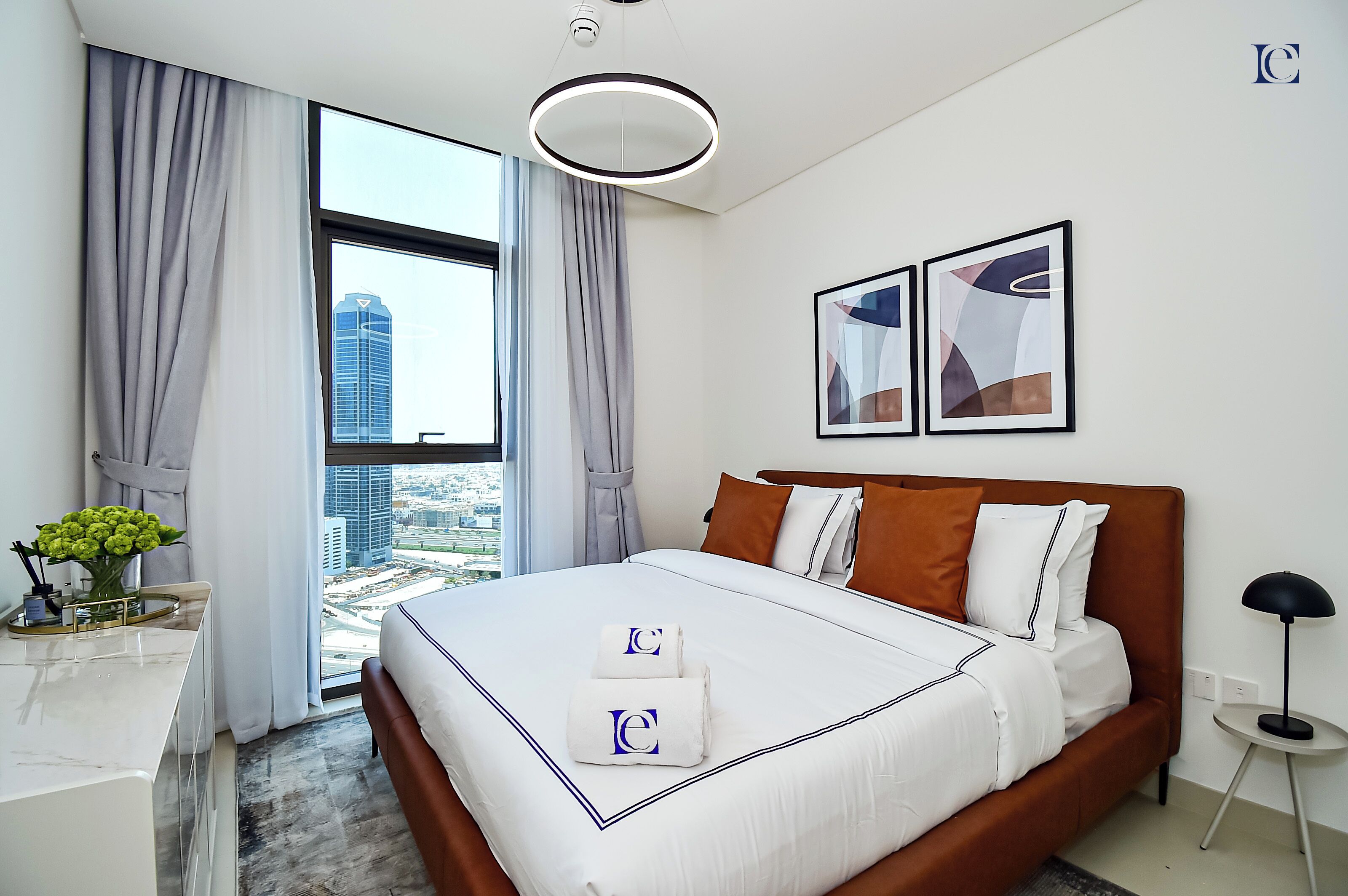 A modern hotel room with a large bed, a desk with a vase of flowers, and a view of a cityscape through the window.