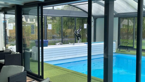 A modern indoor pool area with glass walls, a clear view of the outdoor pool, and a patio with outdoor furniture. - VILLA KEROMER GÎTE DE LO (ETEL)
