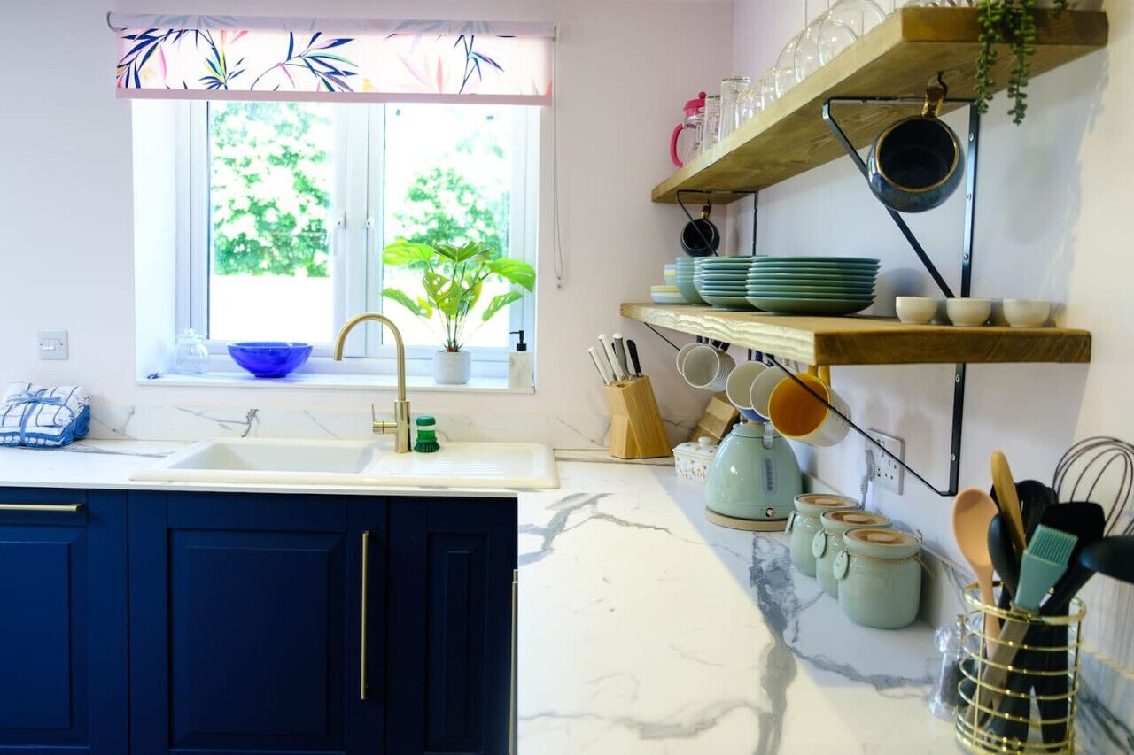 Una cocina con un armario azul, un fregadero, una ventana con una planta y estantes con platos y utensilios.