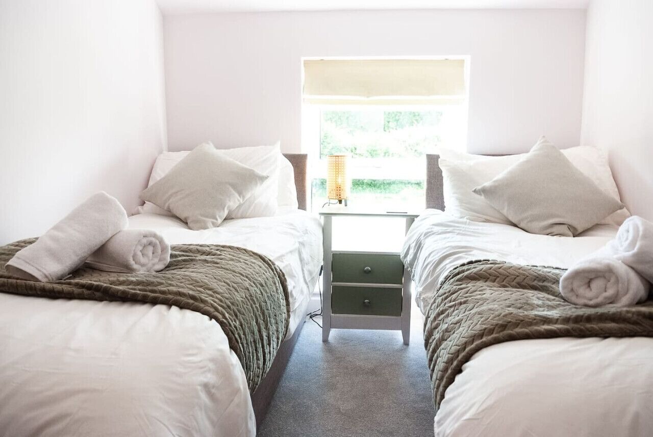 A twin room with two beds, each with a pillow and a rolled towel. There is a nightstand with a lamp between the beds.