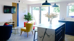A modern living room with a central island, a flat-screen TV mounted on the wall, and a large window providing natural light. - Wave Cottage Bristol (Bristol)