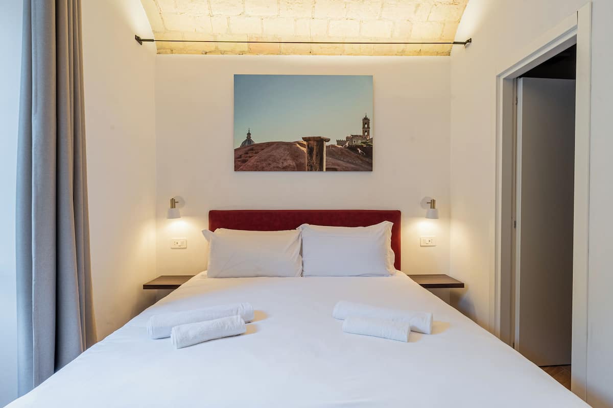 A hotel room with a large bed, white linens, a red headboard, and a painting of a landscape on the wall.