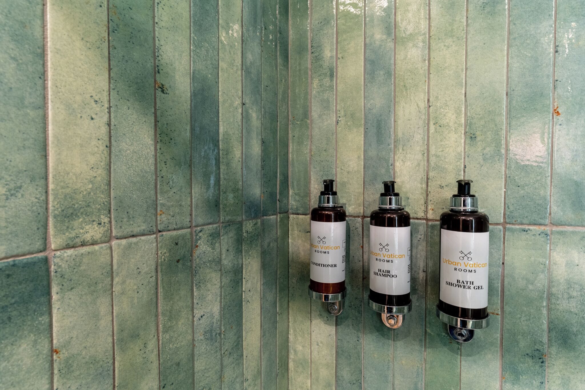 Trois distributeurs de produits d’hygiène, portant l’inscription « Urban Vatican Rooms », sont fixés sur un mur recouvert de carreaux.