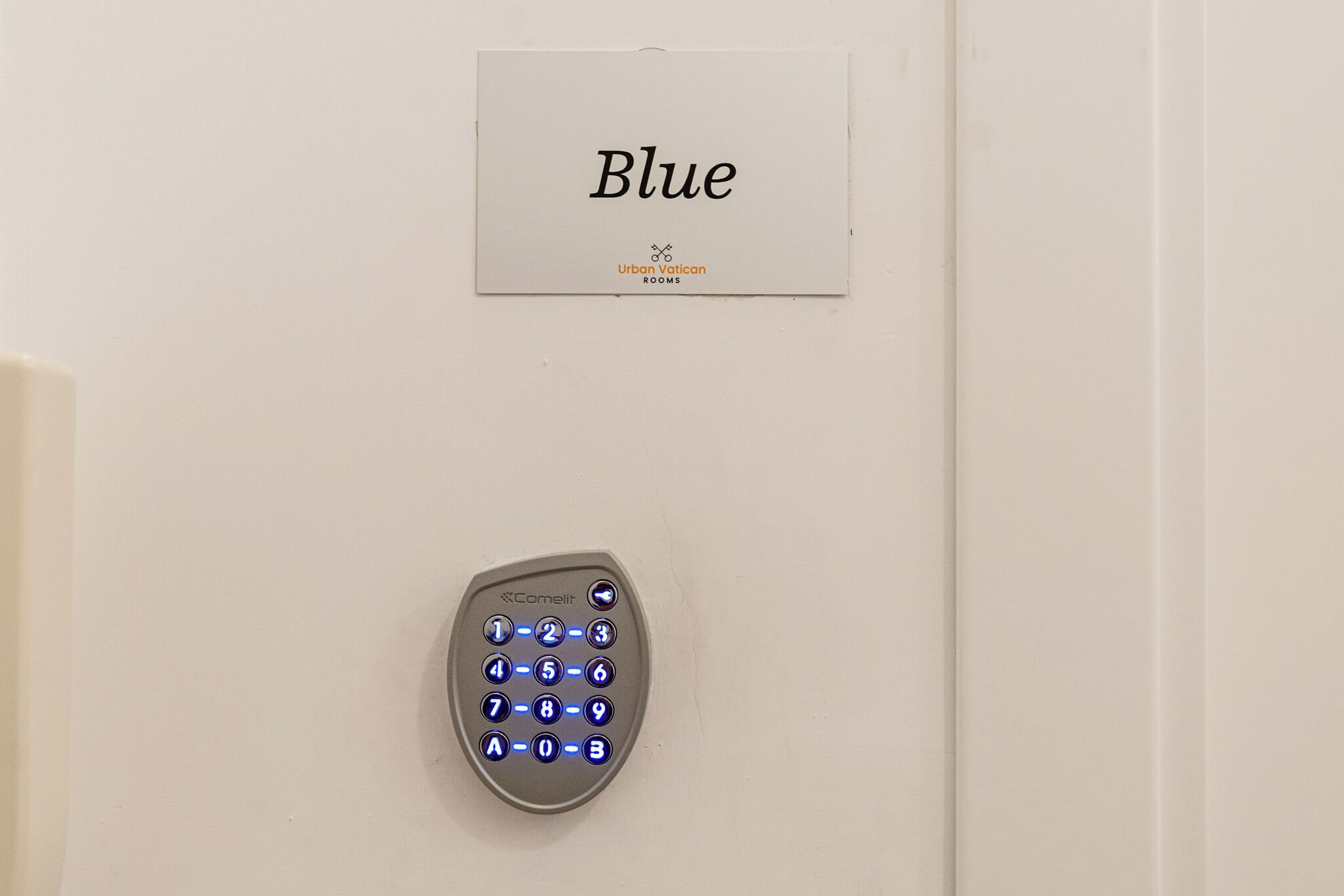 Cerradura electrónica con teclado en una puerta, con un cartel encima que dice 'Blue Urban Vatican ROOMS'.