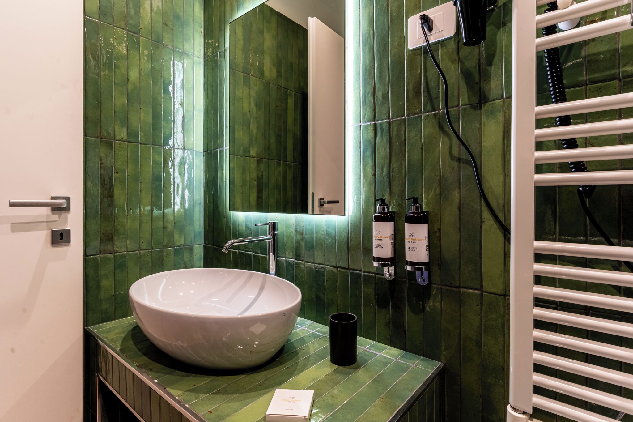 A bathroom with a large white sink, green tiled walls, a mirror, and a towel rack.