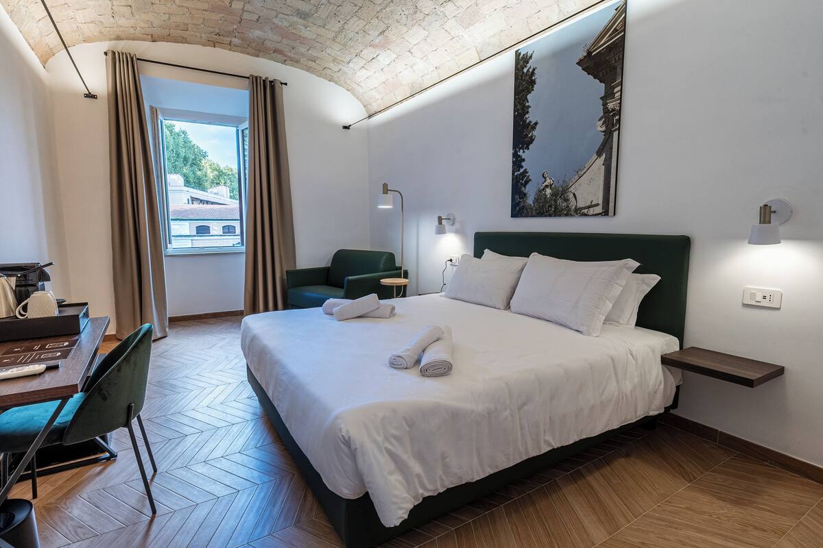 A hotel room with a bed, a desk, a chair, a window with curtains, a painting on the wall, and a lamp.