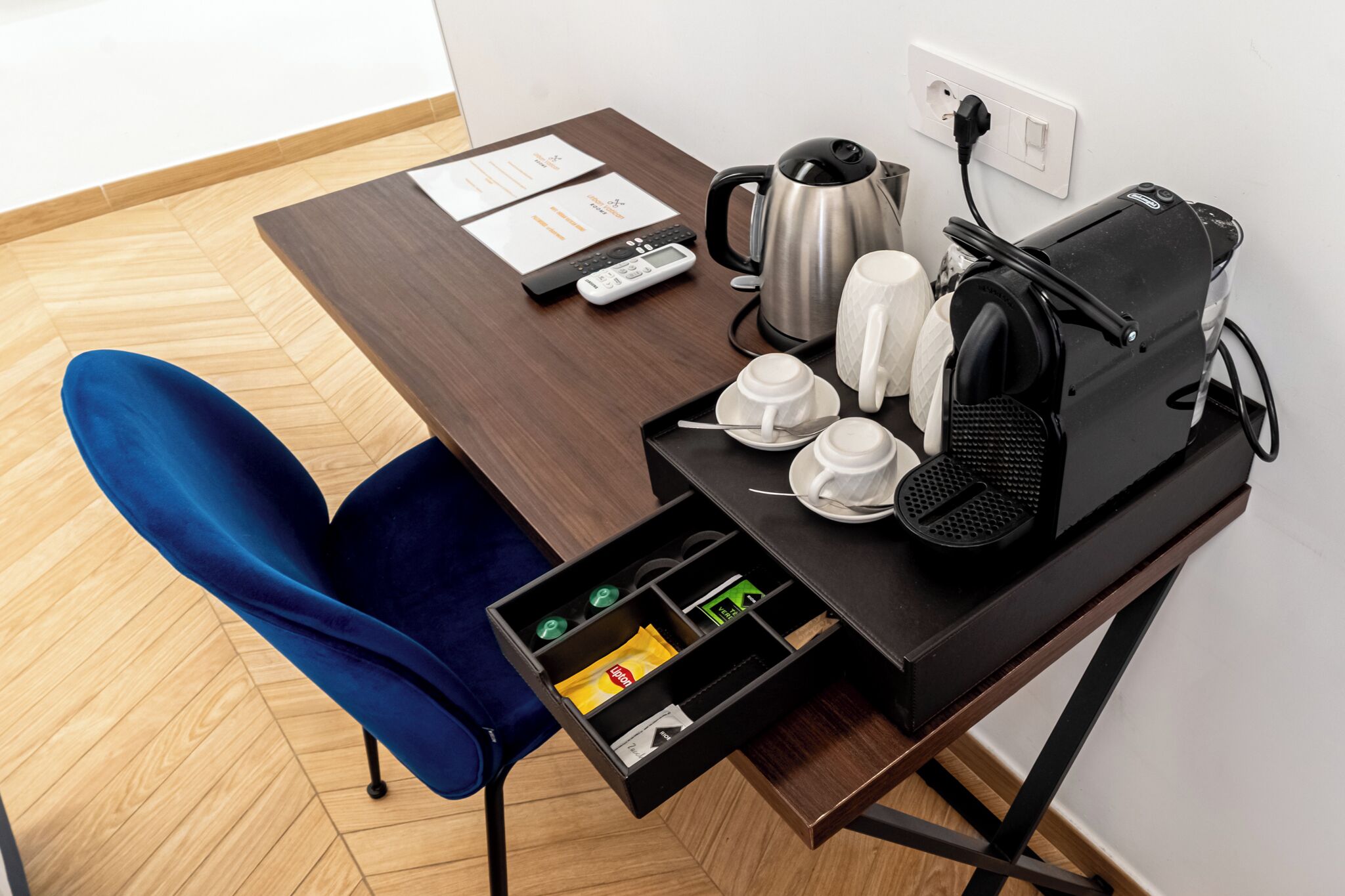 Una habitación de hotel con piso de madera, una silla azul, un escritorio con hervidor, cafetera, parlante y estación de carga.