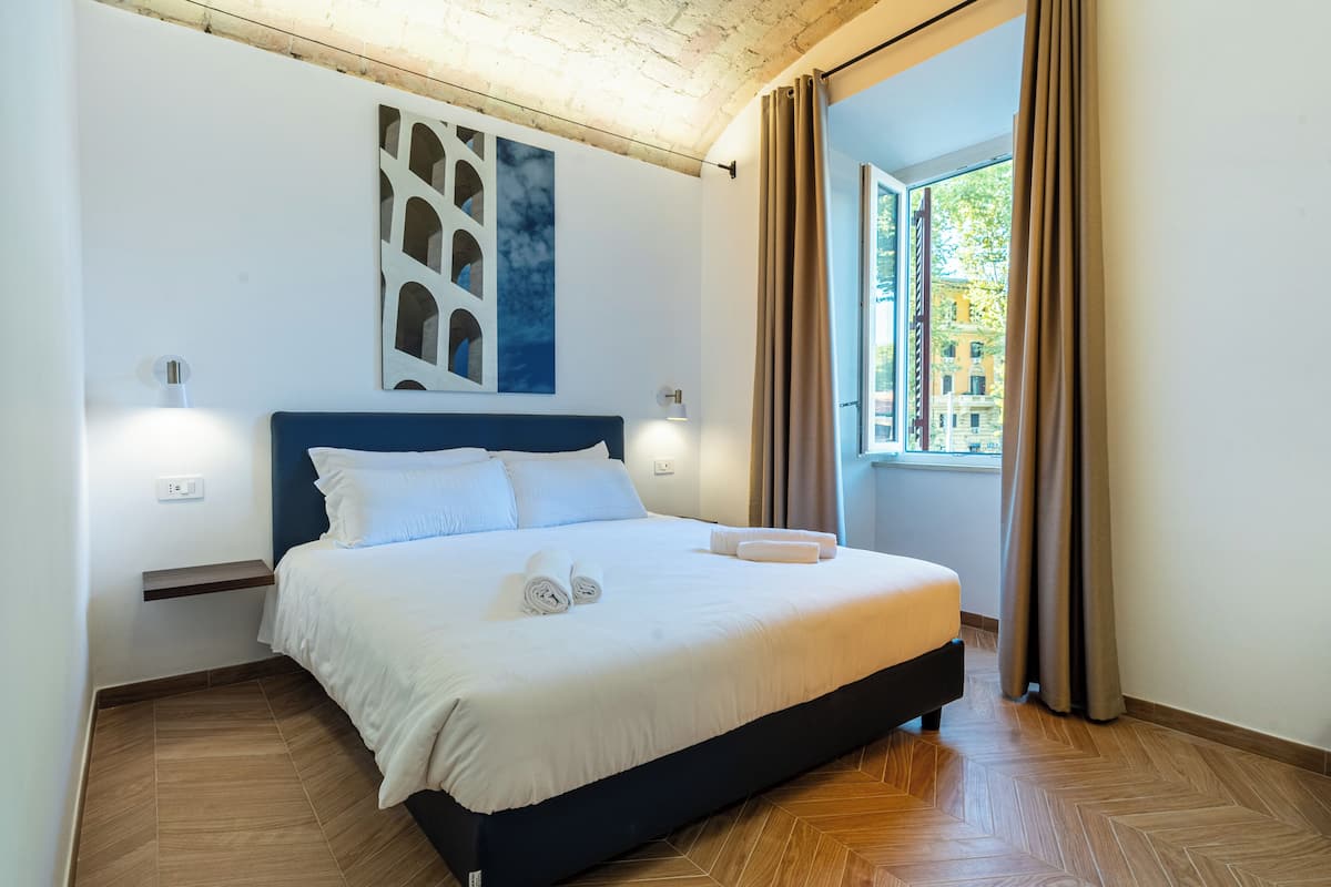 A modern hotel room with a large bed, a headboard with built-in lighting, a wall-mounted artwork, and a view of the city through a window.