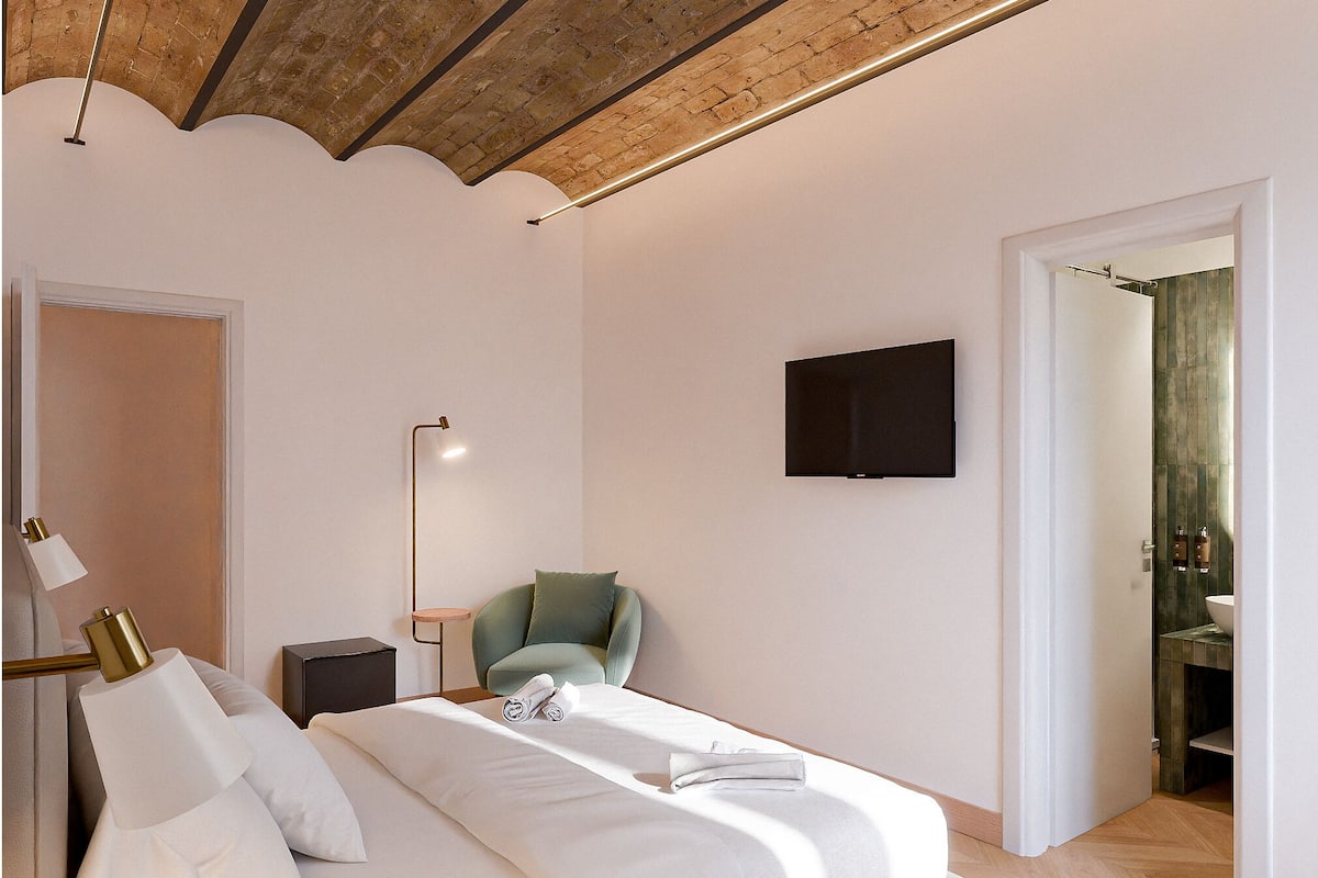 A modern bedroom with a bed, a chair, a TV, and a door leading to a bathroom.