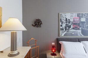 A hotel room with a bed, a nightstand with a lamp, a framed picture on the wall, and a small shelf with figurines.