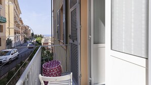 Ένα μπαλκόνι με μια λευκή καρέκλα και ένα τραπέζι με σχέδια λουλουδιών, με θέα σε έναν δρόμο με σταθμευμένα αυτοκίνητα και κτίρια.