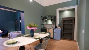 A dining area with a table set for four, a blue chair, a small potted plant, and a kitchenette in the background.