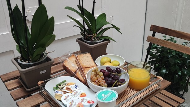 A wooden table with a tray of food, including bread, grapes, and a glass of orange juice, and two potted plants with purple flowers.