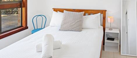 A neatly made bed with white linens and two rolled towels, a wooden headboard, a blue chair, a bedside table with a lamp, and a view of the outdoors through a window.