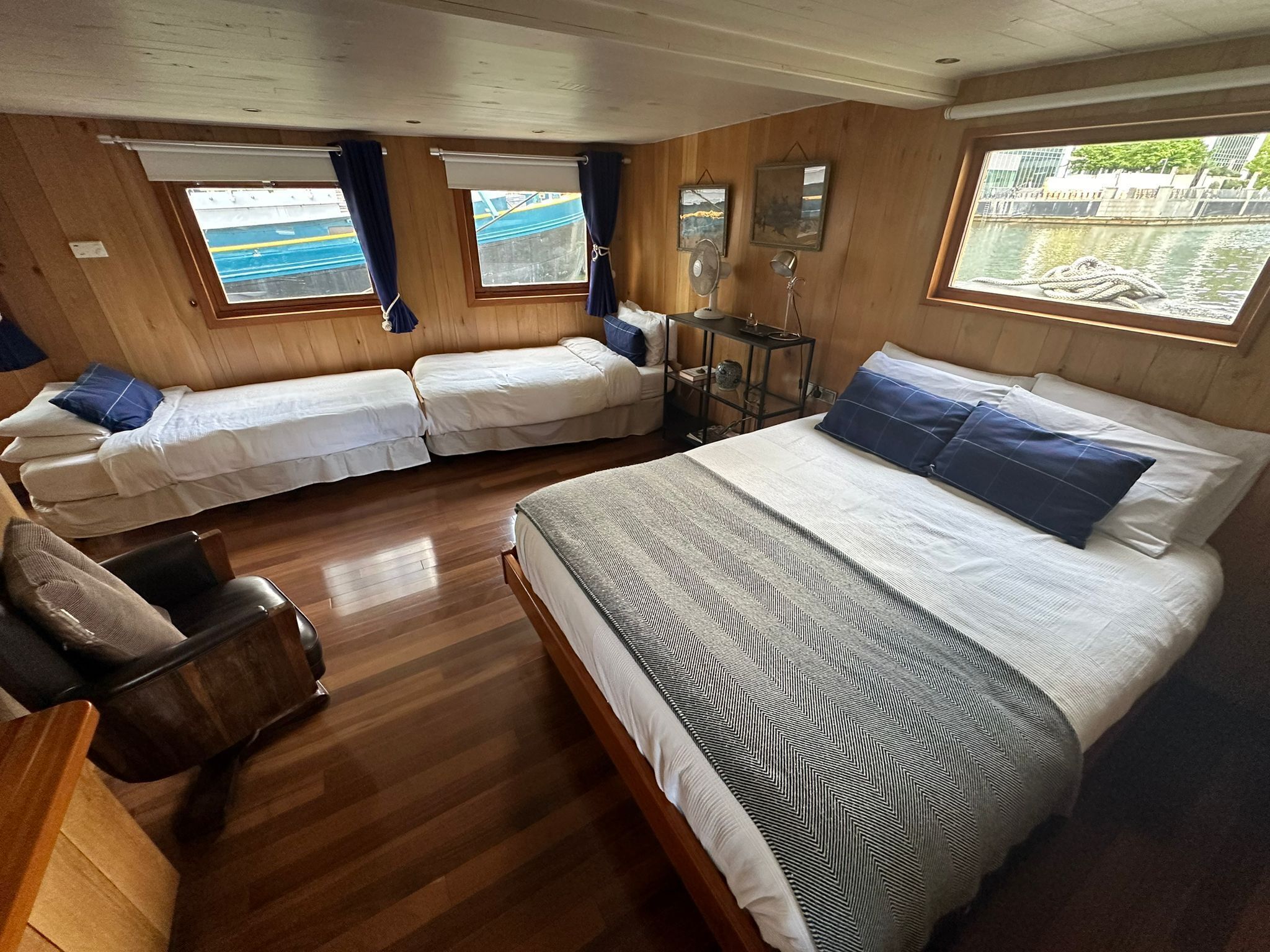 A cabin-style room with two single beds, a larger bed, wooden flooring, and a window with a view of a dock and water.