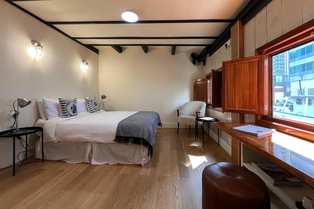 Un dormitorio con cama, mesitas de noche, una silla, un escritorio y una ventana con vistas a edificios.