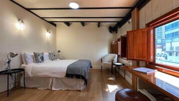 Ein Schlafzimmer mit einem Bett, Nachttischen, einem Sessel, einem Schreibtisch und einem Fenster mit Blick auf Gebäude.