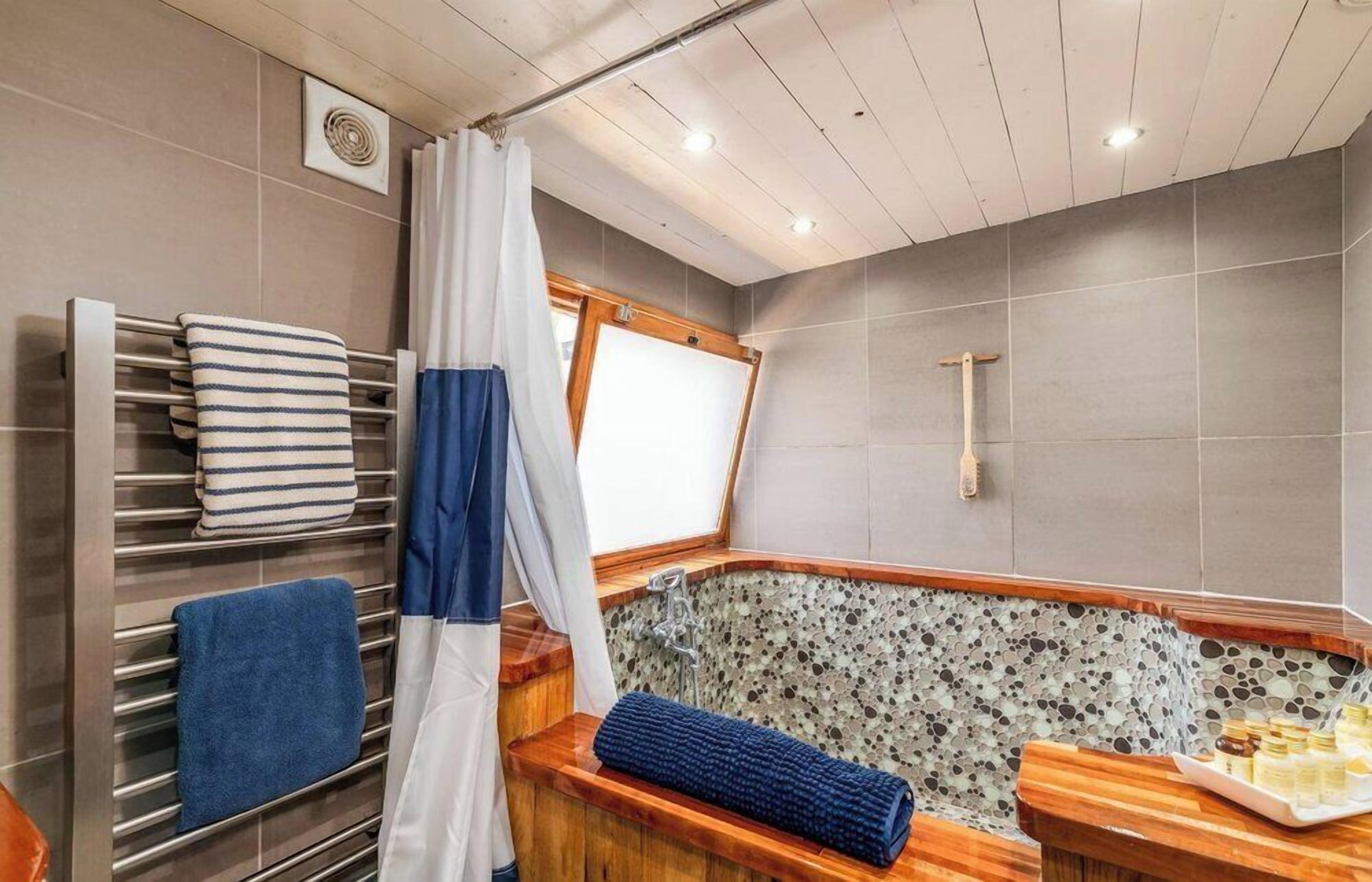 Ein Badezimmer mit einer Badewanne, einem Handtuchhalter mit gestreiften und einfarbigen blauen Handtüchern, einer Holzbank mit einem blauen Handtuch und einer Ablage für Toilettenartikel.