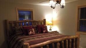 A rustic wooden bed with a headboard, a lamp on a bedside table, and a window with blinds. - The Tug Hill Lodge (Lyons Falls)