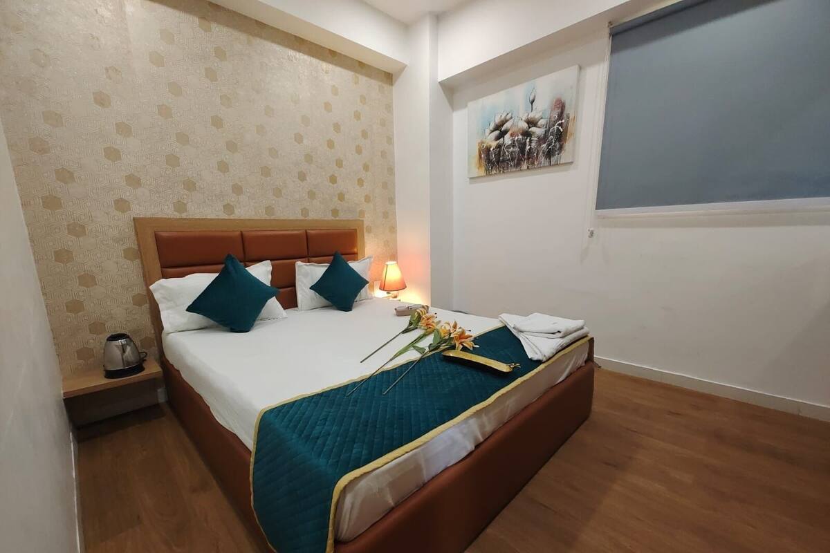 Un lit bien fait, recouvert d’une couverture turquoise, avec une tête de lit en bois et une table de chevet sur laquelle se trouve une bouilloire.