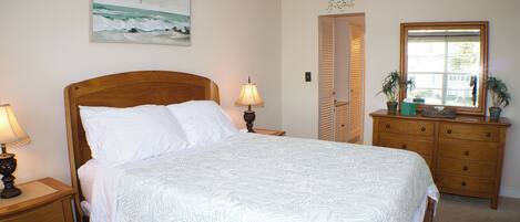 A bedroom with a wooden bed, a nightstand with a lamp, a dresser with two potted plants, and a painting of the ocean on the wall.