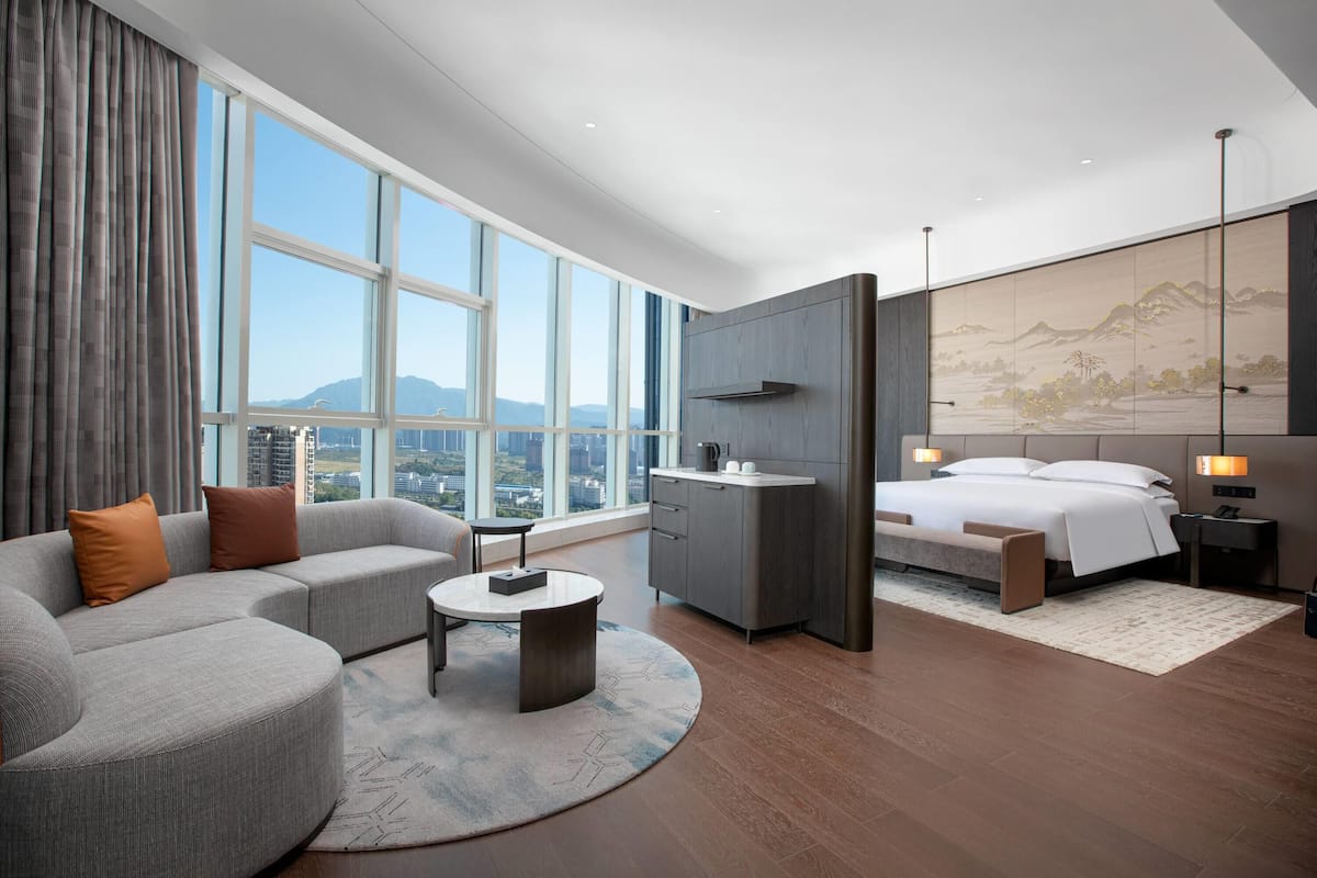 Ein modernes Hotelzimmer mit großem Fenster, einer Couch, einem Bett, einem Schreibtisch und Blick auf die Stadtlandschaft.