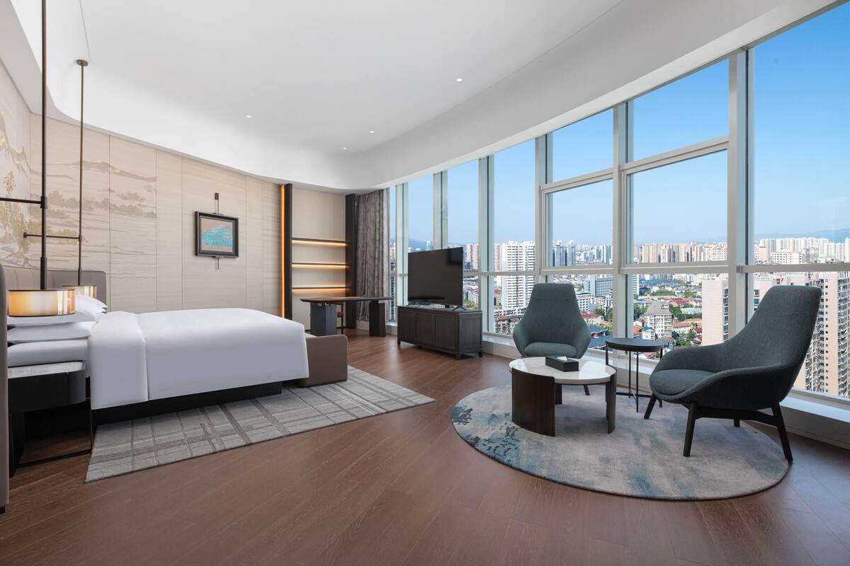 Ein modernes Hotelzimmer mit einem großen Bett, einem Fernseher, einem Schreibtisch und Blick auf die Stadt durch große Fenster.