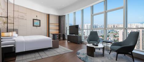A modern hotel room with a large bed, a TV, a desk, and a view of the city through large windows.