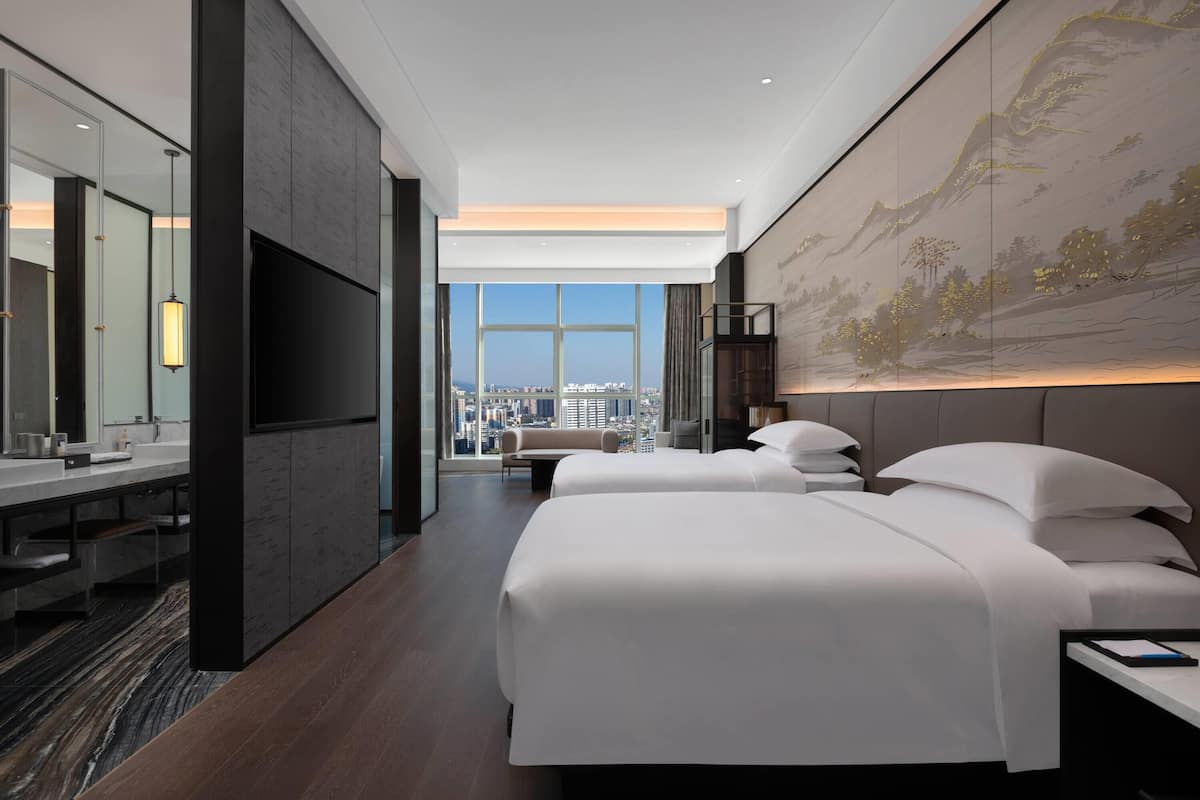 Ein modernes Hotelzimmer mit zwei Betten, einem großen Fenster mit Blick auf die Stadt, einem wandmontierten Fernseher und einem großen Kunstwerk an der Wand.