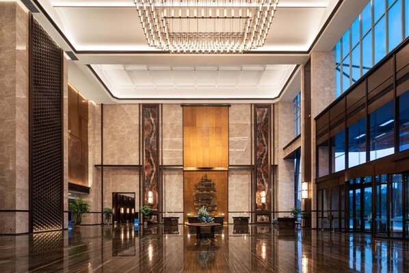 A spacious lobby with a polished marble floor, a central reception desk, and a large chandelier.