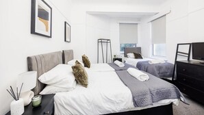 A modern hotel room with two beds, a flat-screen TV, a bedside table with a lamp, and a mirror. - Stylish 2BR Flat | Westcliff Seafront Stay (Westcliff-on-Sea)