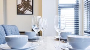 A dining table set with white bowls and wine glasses, a blue upholstered chair, and a window with blinds. - Stylish 2BR Flat | Westcliff Seafront Stay (Westcliff-on-Sea)