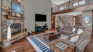 A spacious living room with a stone fireplace, a large sectional sofa, a wooden coffee table, and a staircase leading to an upper level.