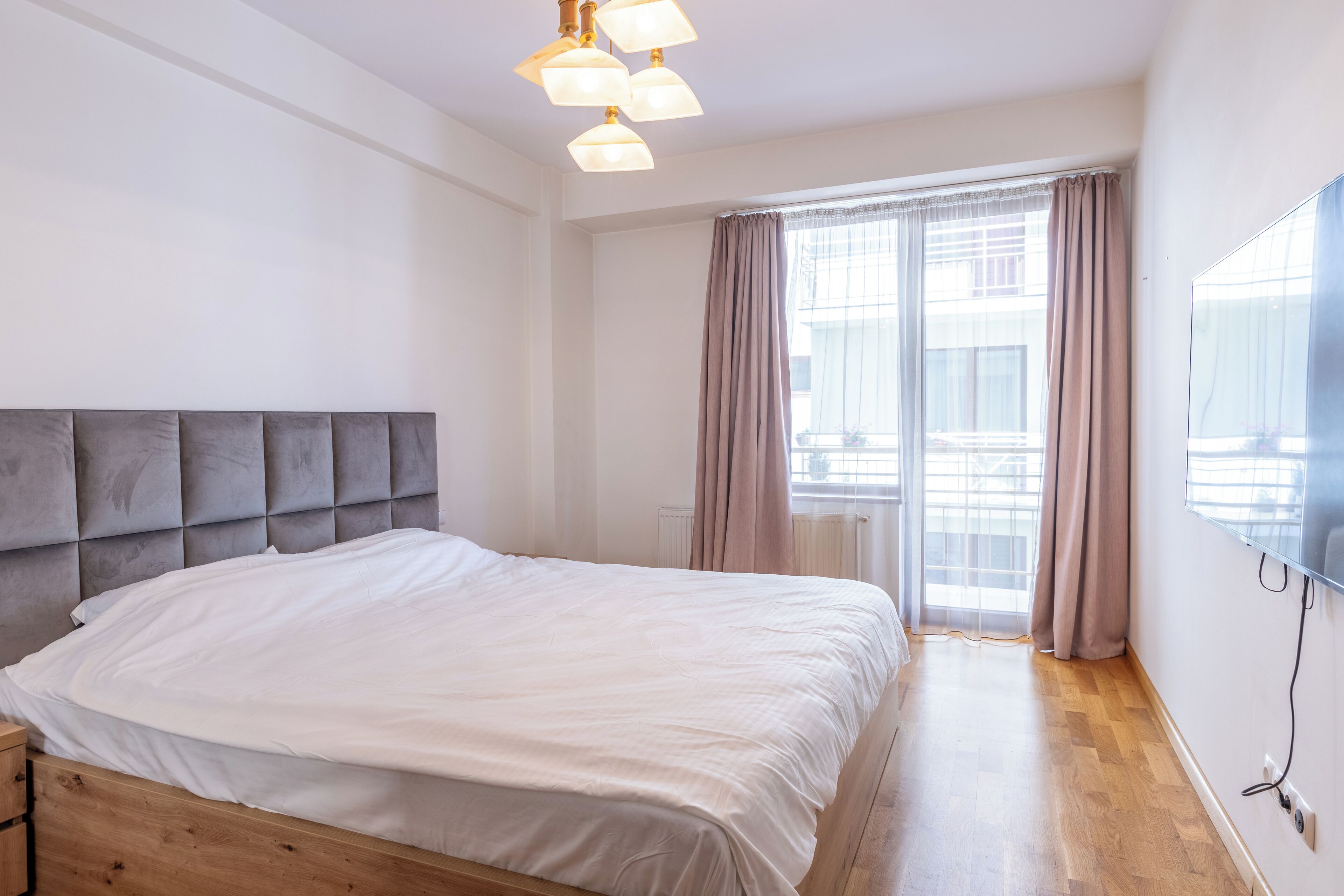 Une chambre avec un grand lit, une tête de lit en bois, un luminaire au plafond, une fenêtre avec des rideaux et un téléviseur fixé au mur.