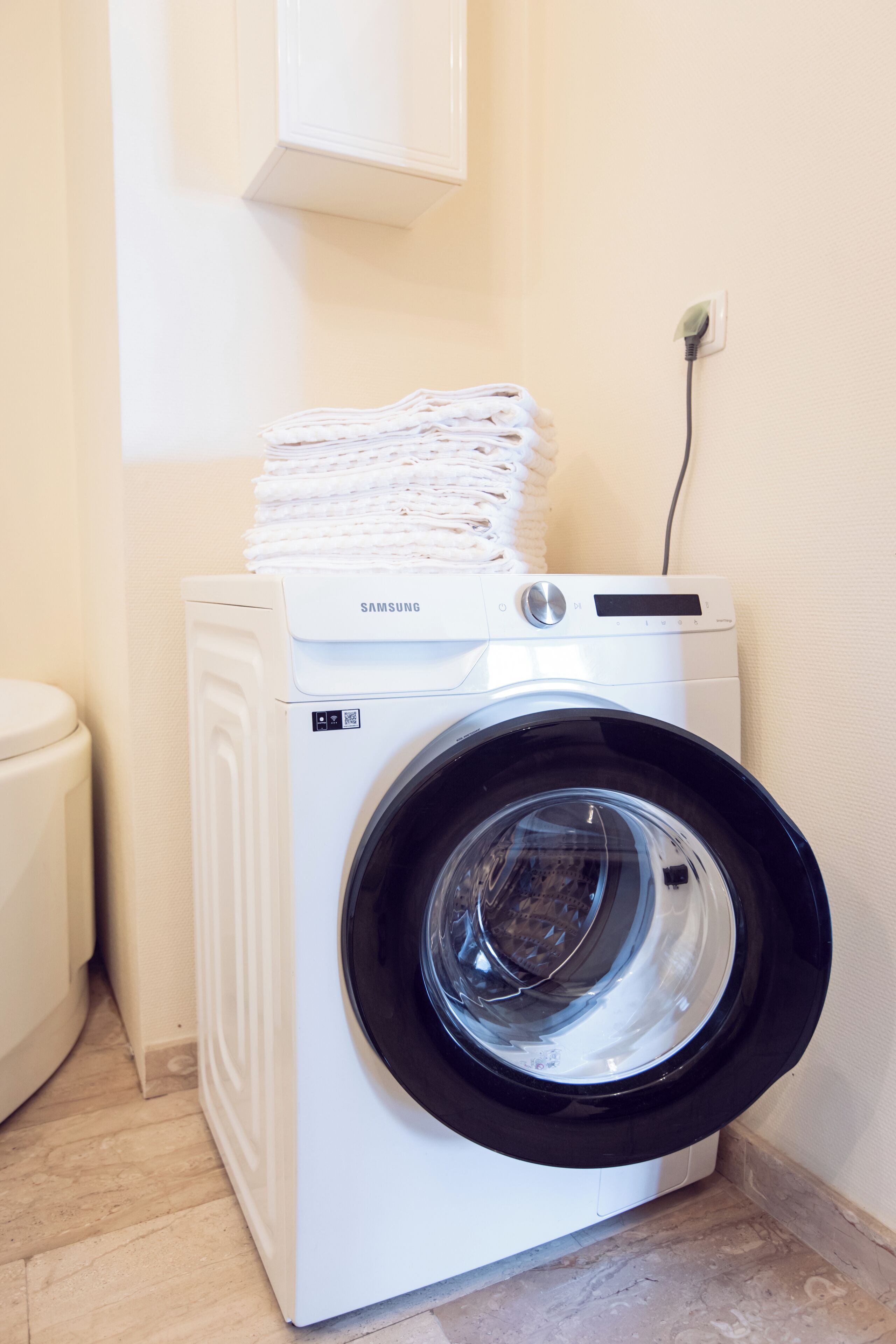 Eine Samsung Waschmaschine mit Frontlader und Glasfront, beladen mit gefalteter Kleidung, in einem Badezimmer mit Toilette und einem fest installierten Steckdosenanschluss.