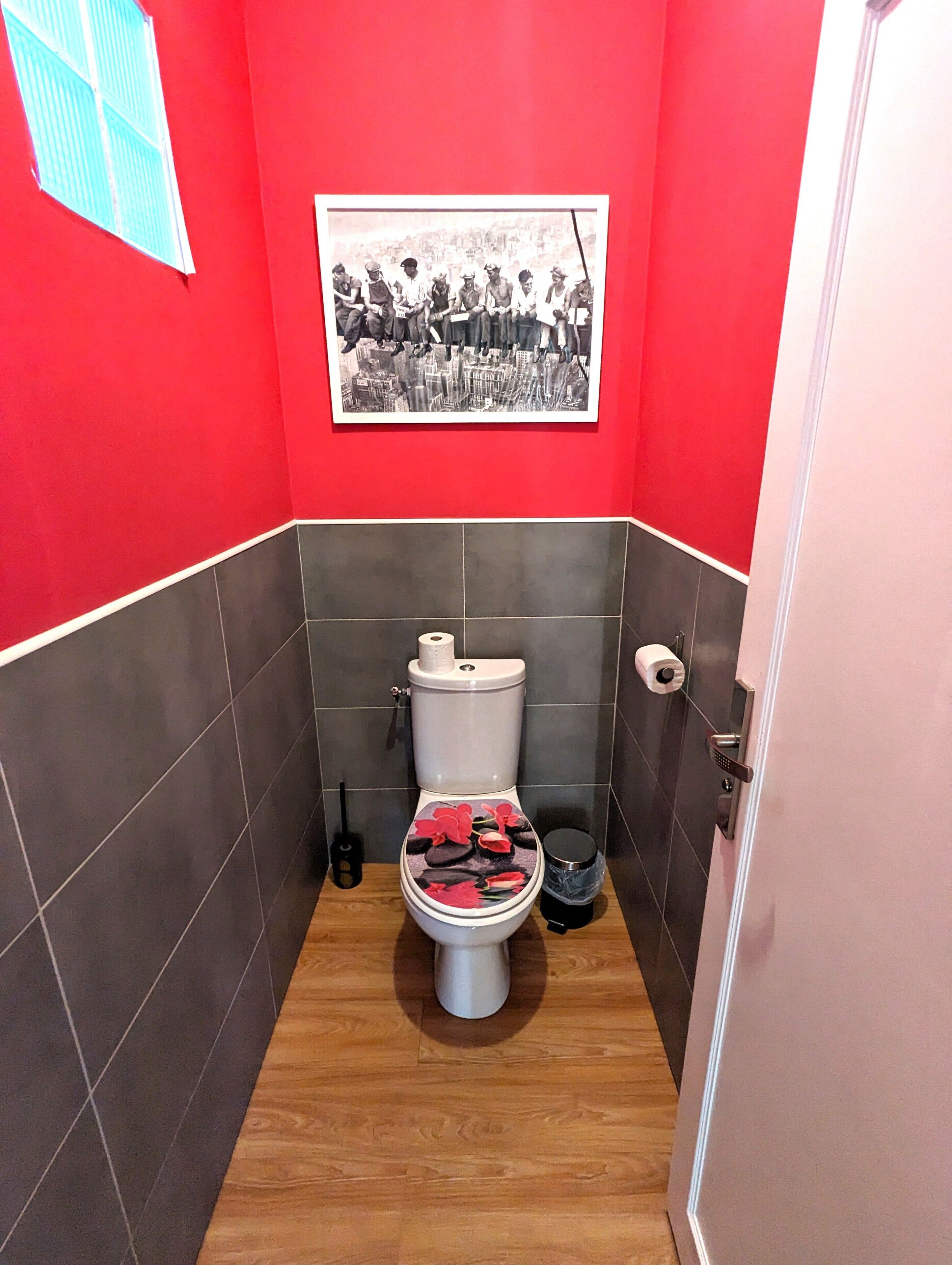 Ein Badezimmer mit roten Wänden, einer Toilette mit floralem Sitz und einem Schwarz-Weiß-Foto an der Wand.