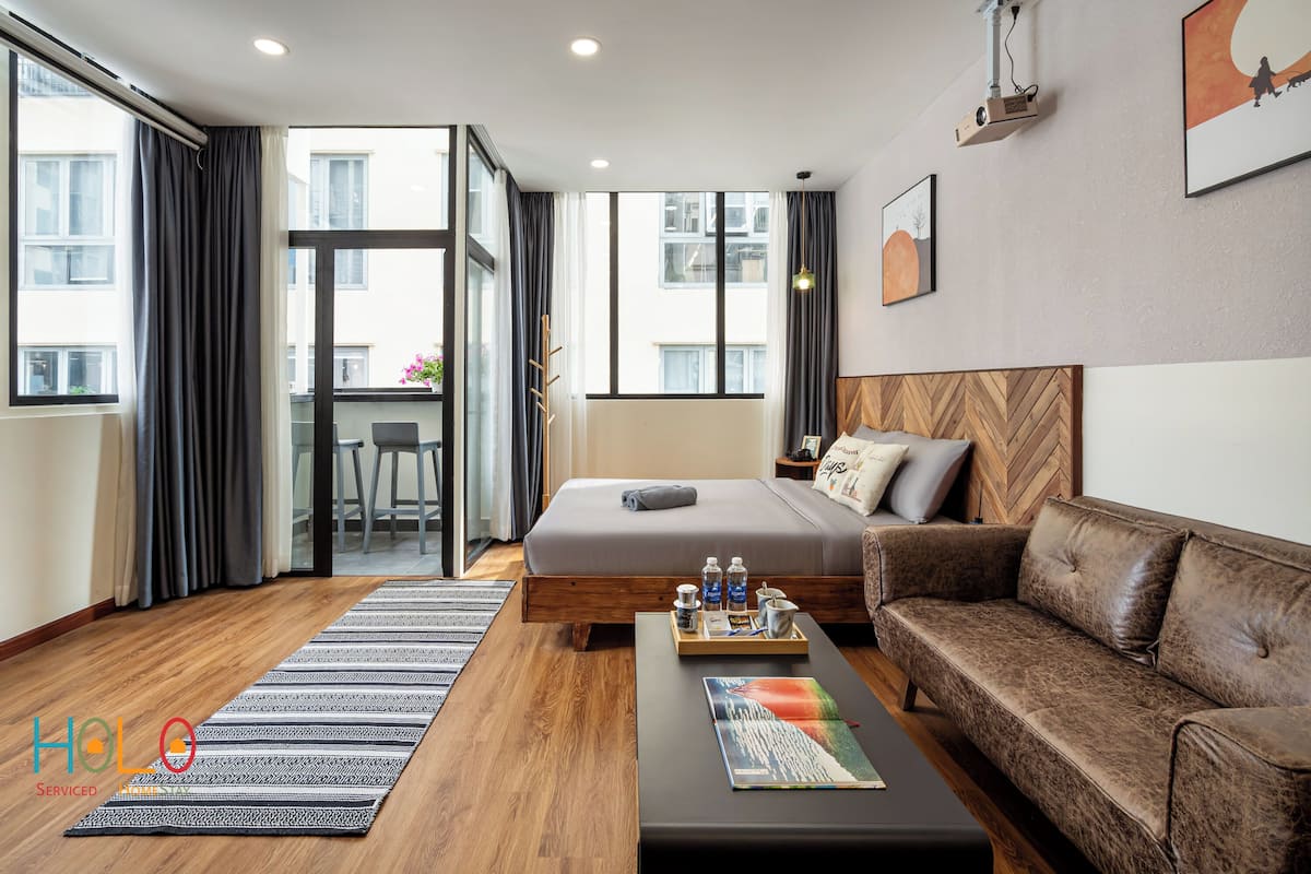 A modern hotel room with a large bed, a leather sofa, a coffee table, and a view of the city through large windows.